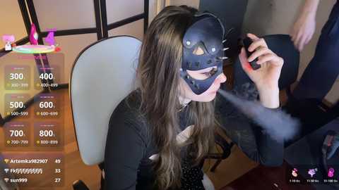 Video of a woman in a black cat mask, sitting on a white chair, holding a cigarette. The background includes a colorful neon sign and a person in a black shirt.