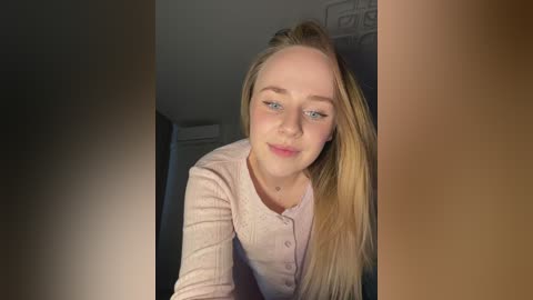 Video of a young, fair-skinned woman with long blonde hair, wearing a light pink ribbed cardigan, smiling softly. The background features a dimly lit, plain room with an air conditioner visible.
