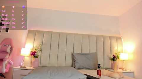 Video of a modern bedroom with a pink gaming chair, white tufted headboard, grey pillows, floral vases, and digital clock.