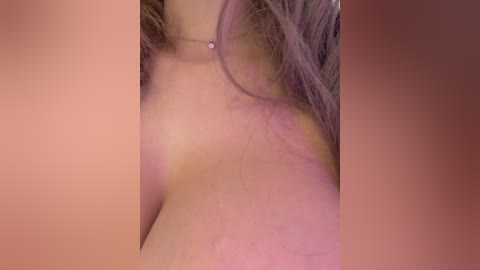 A close-up video of a woman's bare back, showing light skin, shoulder blades, and a delicate silver necklace. Her long, wavy, light brown hair partially covers her neck. The background is blurred, creating a soft, intimate atmosphere.