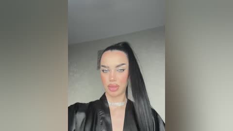 Video of a woman with long black hair in a high ponytail, wearing a black leather jacket and a choker, against a plain, beige wall.