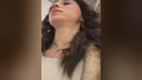 Video of a woman with long, curly brown hair, wearing a white, textured top, looking upward with a serene expression. Background is blurred, creating a soft focus effect.