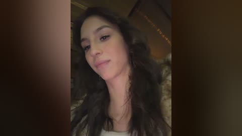 A video of a young woman with long, dark hair and fair skin, wearing a black jacket, smiling slightly. The background features dimly lit, warm-toned indoor lighting and a blurred, textured wall.