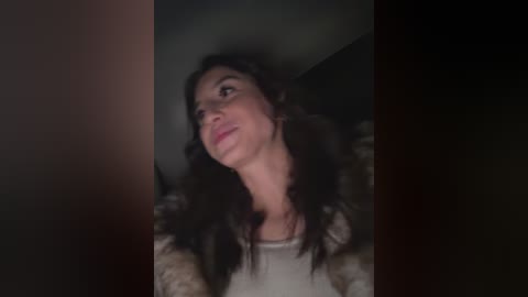 Video of a woman with long, dark hair and a light complexion, wearing a beige sweater, gazing upwards in a dimly lit room with a dark background.