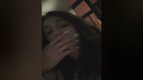 A dimly lit video of a young woman with long, dark hair, eyes closed, and mouth covered by her hand, standing in a dimly lit room with wooden paneling.