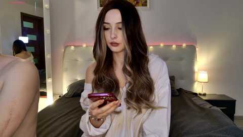 Video of a woman with long, wavy brown hair, wearing an off-shoulder white shirt, sitting on a bed, texting on her red phone in a dimly lit bedroom.