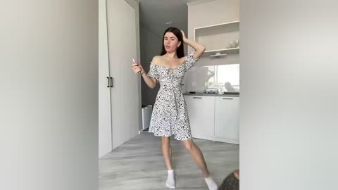 Video of a young woman with fair skin and straight, dark hair in a white, off-shoulder dress, holding a smartphone, in a modern, minimalist kitchen with light wood floors.