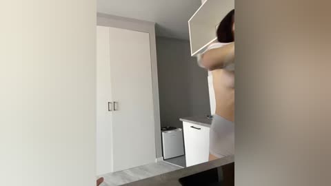 Video of a woman in a modern, minimalist bathroom with light grey walls and white cabinets. She's lifting her shirt, revealing her bare chest. The room has a sleek, contemporary design.
