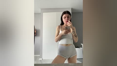 Video of a young woman with fair skin, straight dark hair, and tattoos on her arms, wearing a grey crop top and high-waisted shorts, taking a mirror selfie in a modern, minimalist bathroom with light grey walls and white fixtures.