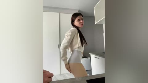 Video of a young woman with long brown hair, wearing a white crop top and high-waisted white shorts, posing in a modern, minimalist kitchen with white cabinets and a dishwasher.