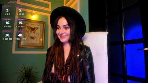 Video of a smiling woman with long brown dreadlocks and a black hat, wearing a leather jacket, in a dimly lit room with green walls and a framed deer picture.