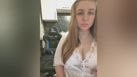 Video of a young Caucasian woman with light skin and long, straight, light brown hair, wearing a white lace nightgown. She sits in a gaming chair in a modern kitchen with white cabinets and a dark granite countertop.