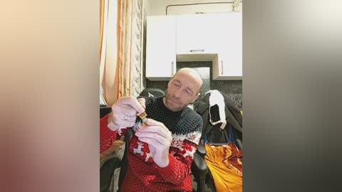Video of a bald, bearded man in a red, white, and black sweater, wearing gloves, seated in a camper van with white cabinets and beige curtains.
