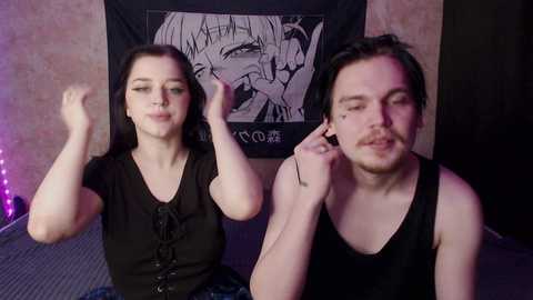 Video of two white people with light skin and dark hair, both wearing black tank tops, sitting on a bed with a black anime poster in the background.