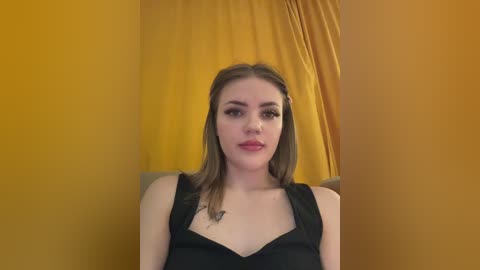 Video of a fair-skinned young woman with long brown hair, wearing a black sleeveless top, against a mustard-yellow curtain backdrop. She has a small butterfly tattoo on her chest.