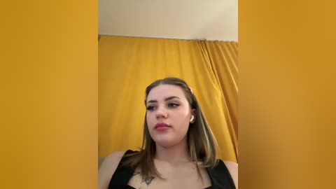 Video of a young Caucasian woman with straight, shoulder-length brown hair, light skin, and wearing black clothing. She has a confident expression, standing against mustard-yellow curtains in a dimly lit room.