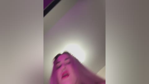 A blurry, low-angle video of a person with short hair, possibly a woman, looking upward, with pinkish light illuminating the ceiling and face.