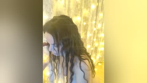 A video of a young woman with long, wavy black hair, wearing a white top, stands in front of a backdrop of glowing, yellow fairy lights. Her face is partially obscured by the lights.