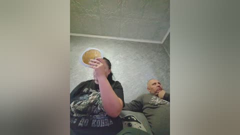 Video of a young woman in a dark graphic t-shirt holding a beer can, sitting next to a bald, older man in a gray sweatshirt, in a dimly lit room with textured, light gray walls.