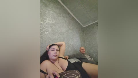 Video of a plus-size woman with dark hair and fair skin in a revealing black outfit lying on a bed, while a bald, older man sits beside her, in a dimly lit room with textured gray walls.
