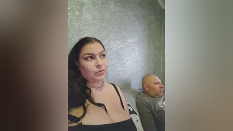 Video of a woman with long black hair, wearing a black dress, looking annoyed, and a bald man in a gray hoodie, both seated on a gray couch against a textured wall.