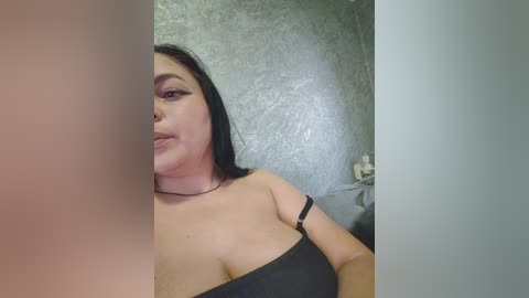 Video of a curvy woman with long black hair, wearing a black strapless top, smiling slightly. Background shows a textured gray wall and a partially visible white door.