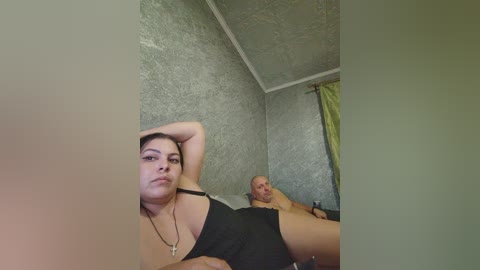 Video of a young woman with light skin and dark hair, wearing a black dress, lying next to a bald man in a gray room.
