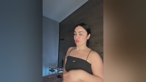 Video of a woman with light skin, dark hair, and medium build, wearing a black crop top, sitting on a bed with a wooden headboard.