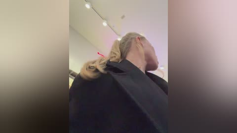 Video of a blonde woman with a ponytail, wearing black hairdressing robes, standing in a salon with overhead lights.