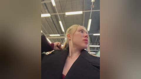 Video of a young blonde woman in a black blazer, adjusting her hair in an industrial setting with metallic ceilings and fluorescent lights.