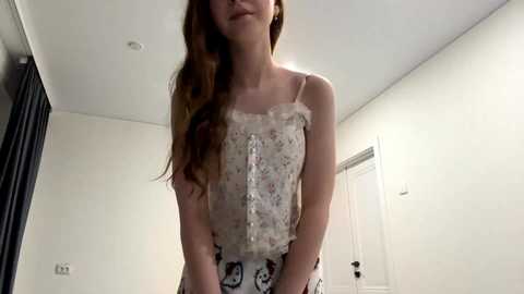 Video of a young Caucasian woman with long auburn hair, wearing a floral-patterned sleeveless top and high-waisted pants. She stands in a minimalist, well-lit room with white walls, a door, and a black curtain.