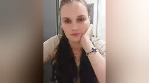 Video of a fair-skinned woman with blonde braided hair, resting her head on her hand, wearing a beige top and black scarf, in a bathroom with a tiled wall and a partially visible mirror.