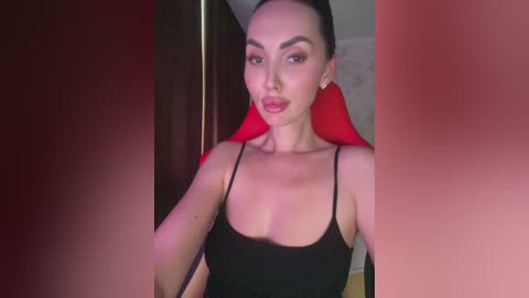 Video of a light-skinned woman with dark hair, wearing a black tank top, sitting in a red chair. Background is dimly lit, with blurry dark and red tones.