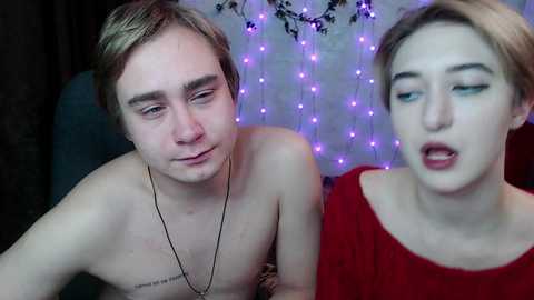 Video of a shirtless, slender, light-skinned man with short brown hair and a necklace, and a fair-skinned woman with blonde hair and red lipstick, both wearing casual attire, indoors with purple string lights and floral decor.