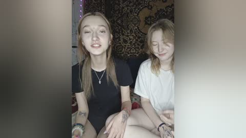 Video of two young women with pale skin, sitting together indoors. One with long brown hair wears a black top, the other with blonde hair in a white t-shirt. Both have tattoos. Background includes a patterned tapestry.
