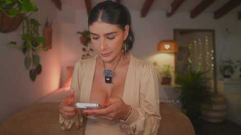 Video of a woman with black hair, medium breasts, wearing a beige dress, playing a smartphone, in a cozy, warmly lit room with potted plants and wooden beams.