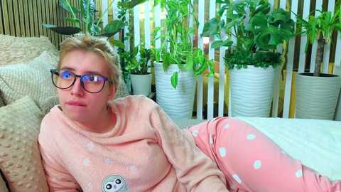 Video of a fair-skinned woman with short blonde hair and glasses, wearing a peach sweatshirt and polka-dot pajama pants, lying on a beige couch with potted plants in the background.