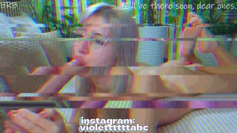 A blurry, distorted video screenshot of a young, blonde woman with glasses, lying on her stomach, performing oral sex on a man. Text overlay: \"ill be there soon, dear ones,\" and \"instagram: violerettitab.\