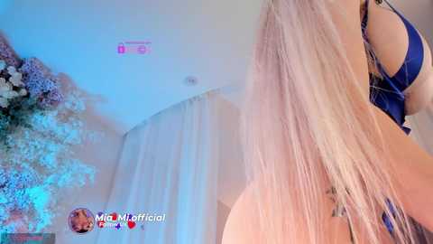 A video of a blonde woman in a dark blue lingerie outfit, taken from a low angle, showcasing her long hair and curves. The background features a floral arrangement and white curtains.