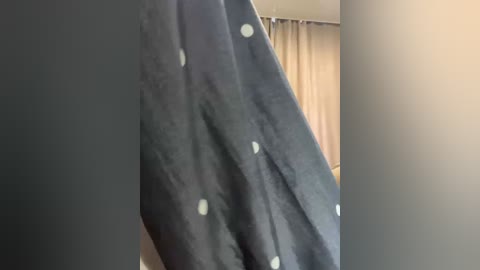 A video of a person in a dark navy blue robe with white polka dots, standing in a room with beige walls and a beige curtain. The image is slightly blurry.