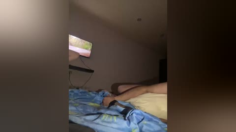Video of a dimly lit bedroom with a flat-screen TV mounted on a wall, displaying a greenish image. A person, possibly male, is lying on a bed with blue and yellow bedding, legs extended.