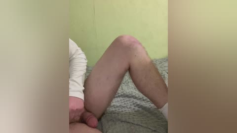 A video showing a person's lower body, with light skin and visible knee, lying on a grey knitted blanket in a room with green walls. The person's legs are bent, and the hand of another person is visible, grasping the blanket.
