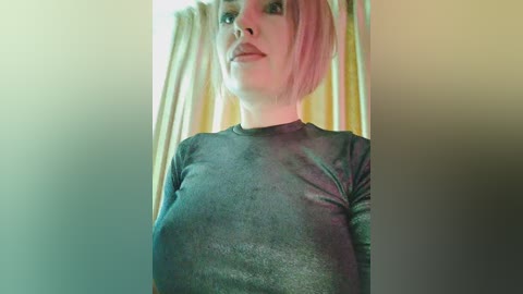 Video of a fair-skinned woman with short pink hair and a black, form-fitting top, standing in a room with yellow curtains and soft lighting.