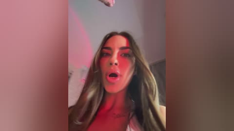 A video of a young woman with long brown hair, wearing a white top, standing in a dimly lit room, her mouth open in an expression of surprise or shock.