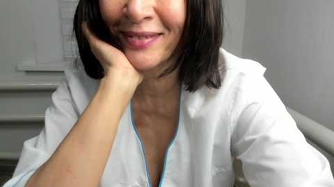 Video of an East Asian woman with shoulder-length black hair, wearing a white lab coat with light blue trim, smiling while resting her chin on her hand in a brightly lit, clinical setting.