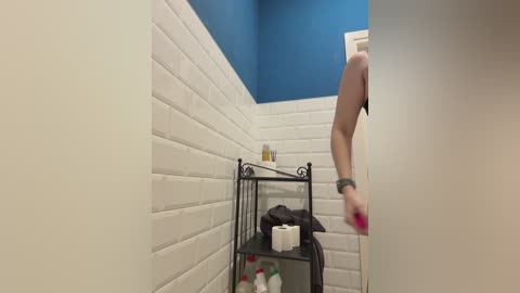 A video of a bathroom with white brick walls, a blue ceiling, and a metal shelf holding toiletries. A person, partially visible, wearing white high heels is seen from the waist down.