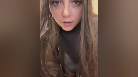 A video of a young woman with long, wavy brown hair, light skin, and brown eyes, wearing a black turtleneck sweater, leaning forward with a serious expression.