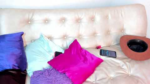 A video of a plush, white, tufted leather sofa with a bright pink pillow and a blue one. A brown cowboy hat, a remote control, and a purple blanket are scattered on the cushions.