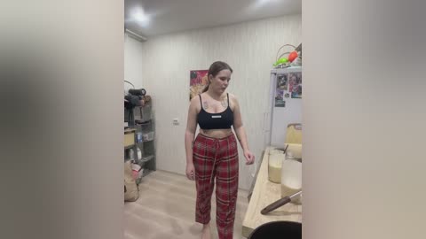 A video of a young woman in a minimalist kitchen with white walls, wearing a black sports bra and red plaid pants.