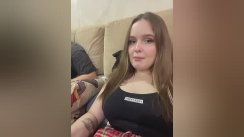Video of a young woman with long brown hair, wearing a black tank top with a white label, sitting on a beige couch, partially covered by a red and white plaid blanket.
